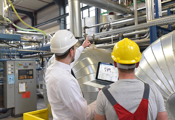 Zwei Männer mit Helm und Laptop in der Hand an Maschine